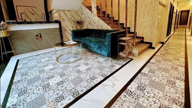 A modern hotel lobby with a marble reception desk, a blue sofa, a staircase with wooden railings, and patterned carpet. - Rich Hotel (Mumbai)