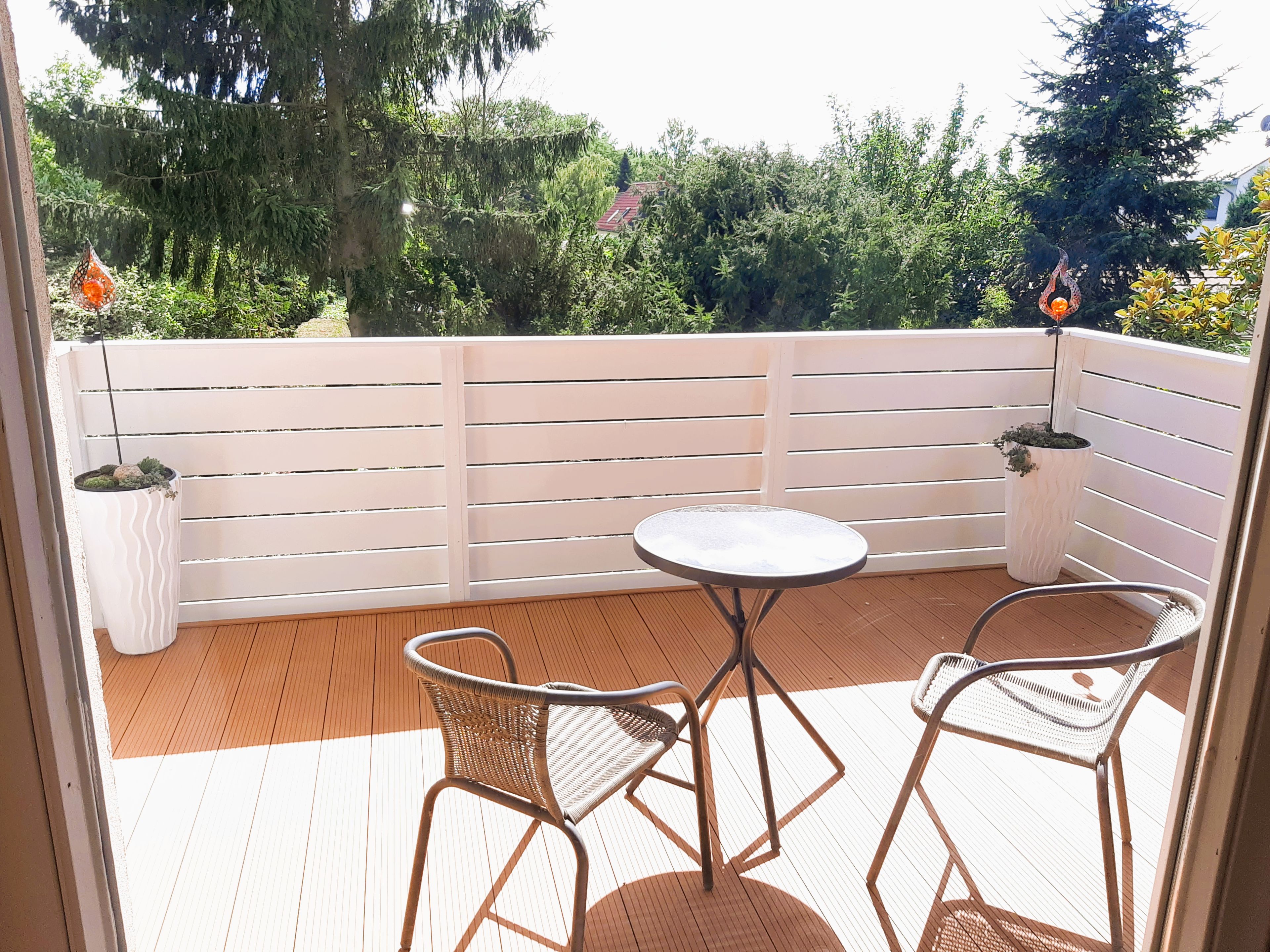 Ein Balkon mit Holzboden, weißer Reling und zwei Korbstühlen um einen kleinen runden Tisch.
