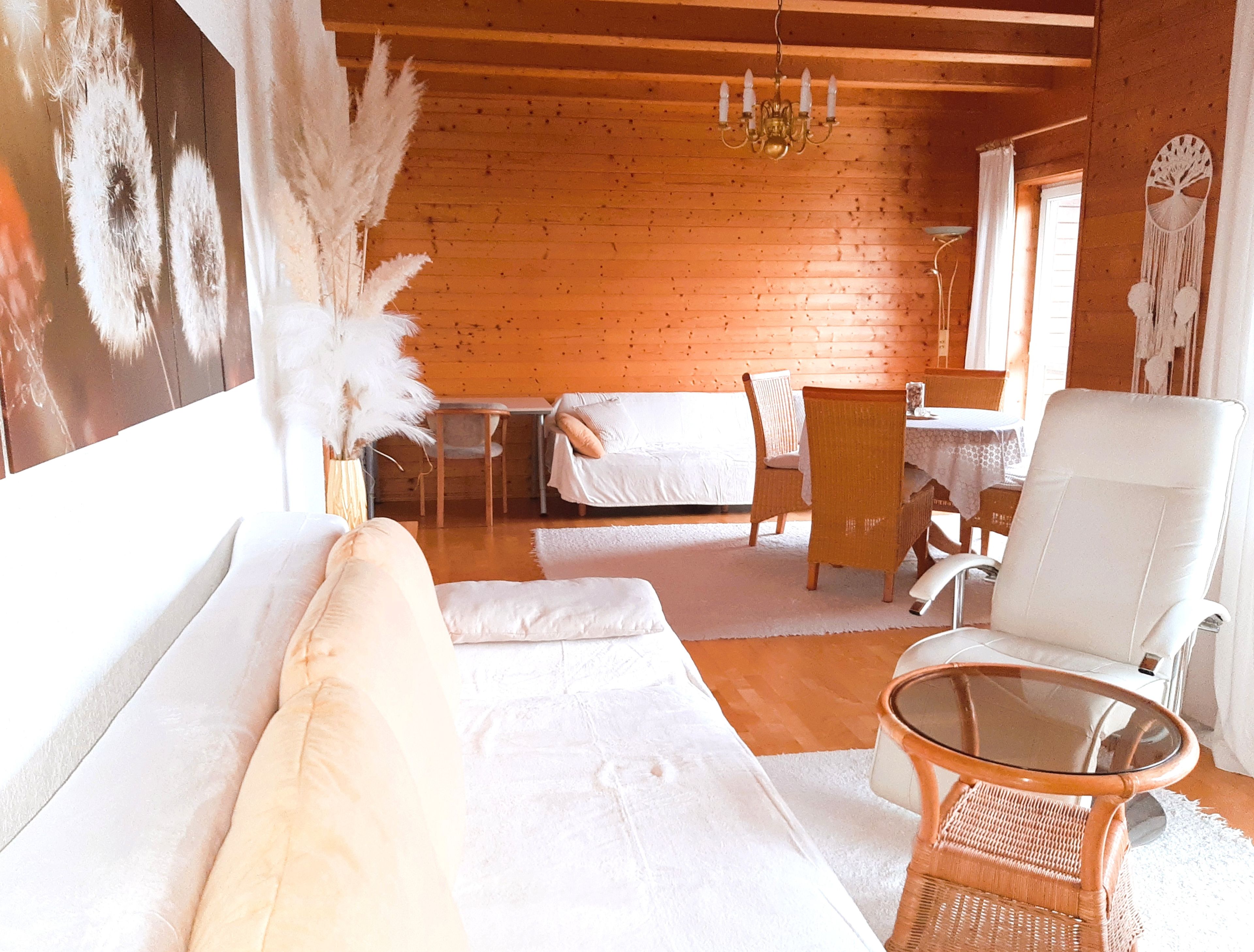 Een knus ingerichte kamer met een houten plafond, een witte bank, een rieten stoel, een glazen tafel en een bed met witte lakens.