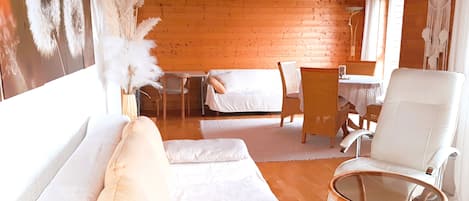 A cozy room with a wooden ceiling, a white sofa, a wicker chair, a glass table, and a bed with white bedding.