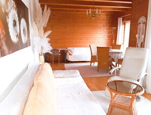A cozy room with a wooden ceiling, a white sofa, a wicker chair, a glass table, and a bed with white bedding.