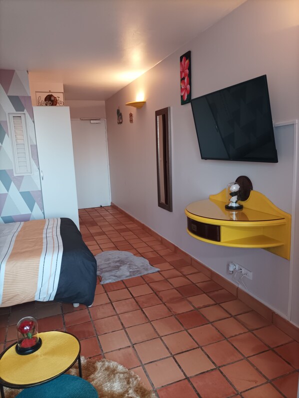 A bedroom with a bed, a yellow bedside table, a TV mounted on the wall, and a tiled floor. - STUDIO ROUGE GORGE IN TARTANE (Tartane)