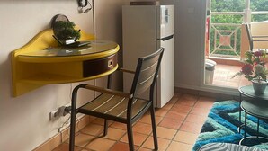 Una habitación compacta con una televisión montada en la pared, un refrigerador pequeño, una mesa amarilla con una silla y una ventana con vistas a zonas verdes.