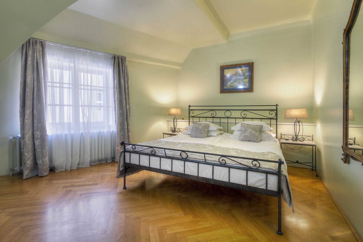 A bedroom with a metal bed frame, two bedside tables with lamps, a large window with curtains, and a framed picture on the wall.