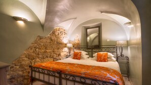 A bedroom with a stone wall, a bed with an orange bedspread, a bedside table with a lamp, and a mirror on the wall. - Black Star Suites (Prague)