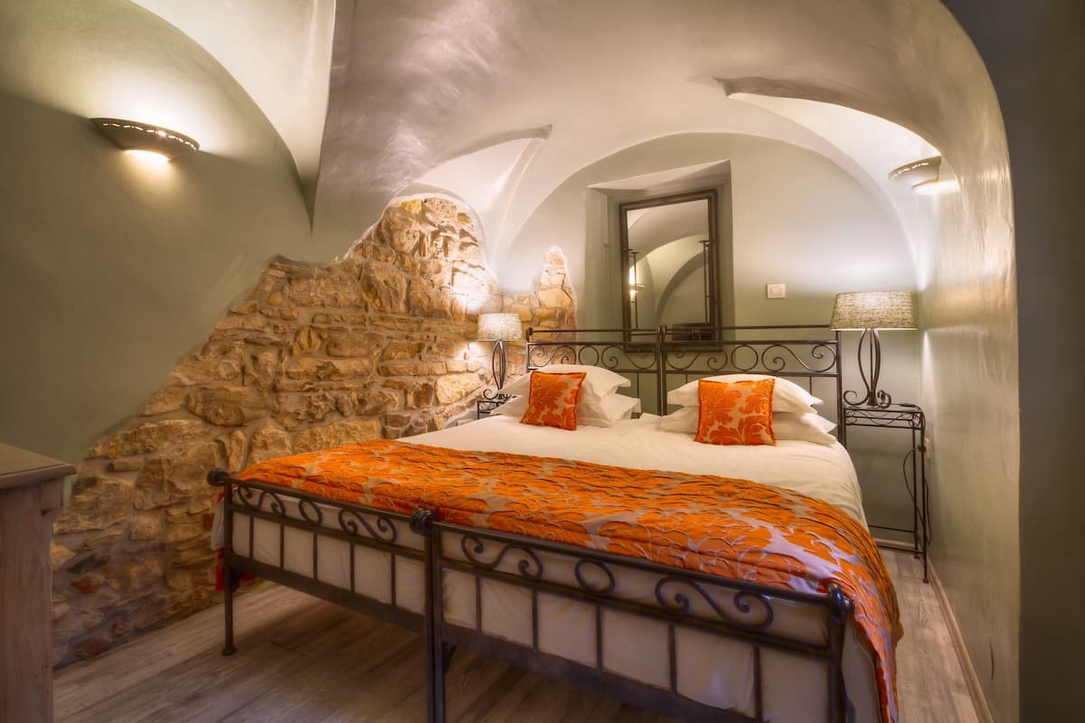 A bedroom with a stone wall, a bed with an orange bedspread, a bedside table with a lamp, and a mirror on the wall.