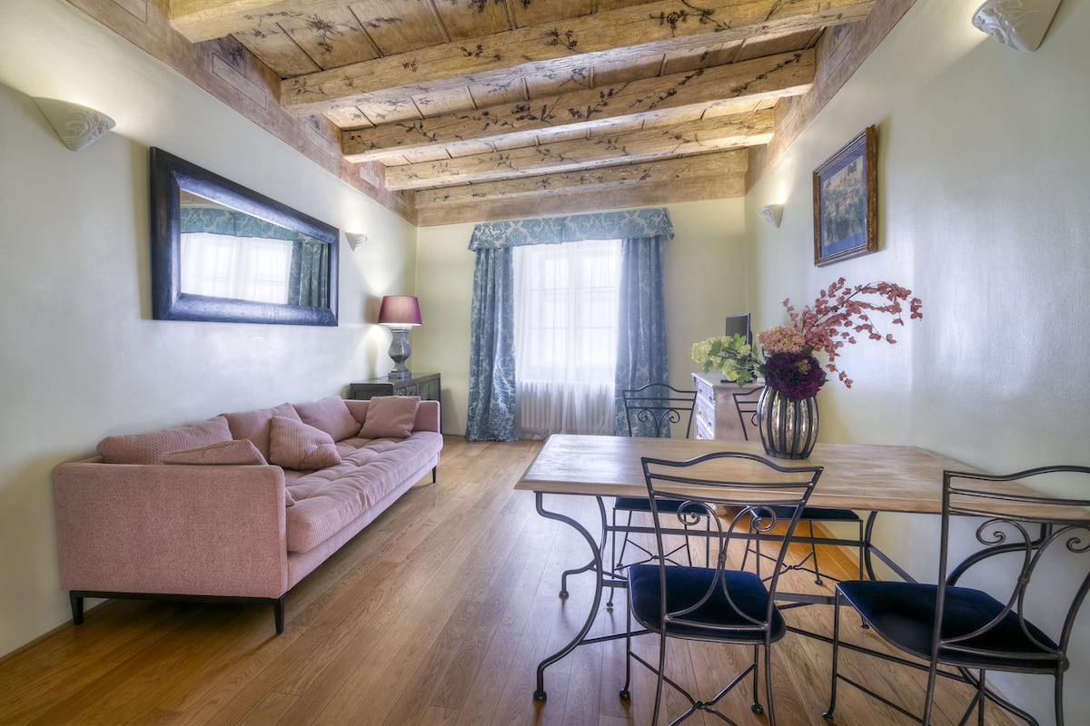 A cozy living room with a wooden ceiling, a pink sofa, a dining table with metal chairs, and a vase of flowers.