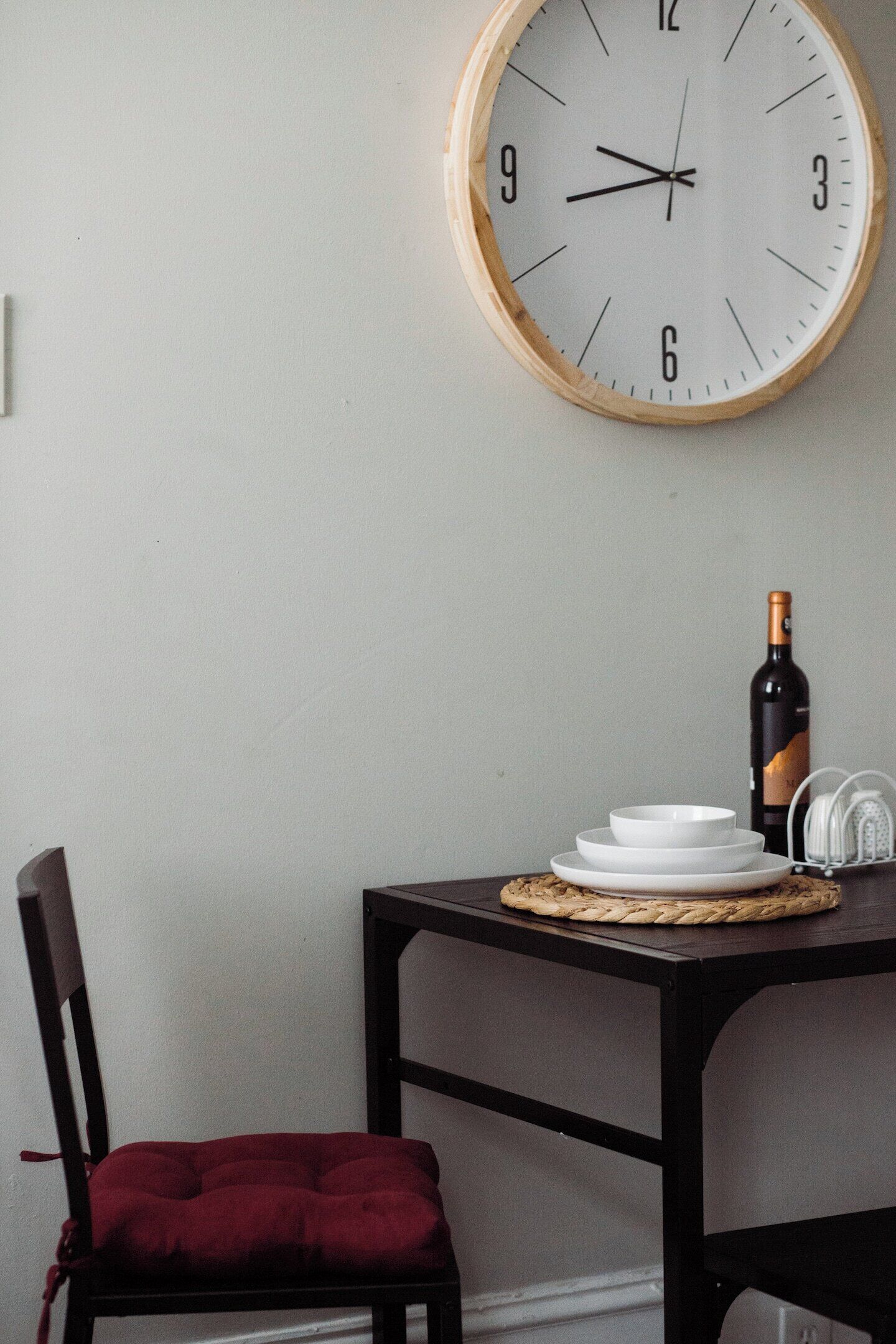 A dining area with a wooden table, a red cushioned chair, a wall-mounted clock, and a bottle of wine.
