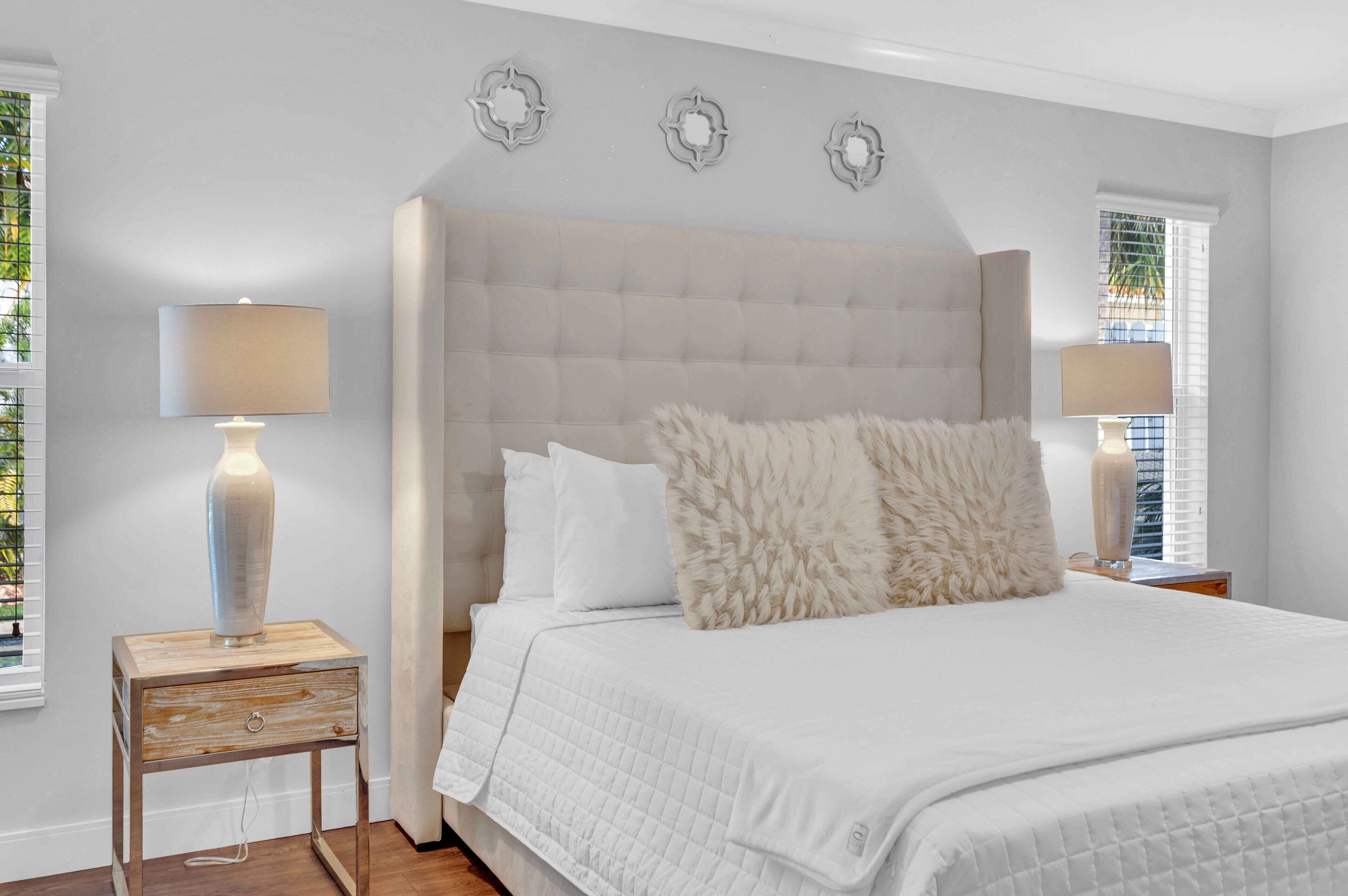 Une chambre à coucher avec une tête de lit capitonnée, du linge de lit blanc, une table de chevet en bois avec une lampe et des œuvres d’art décoratives au mur.