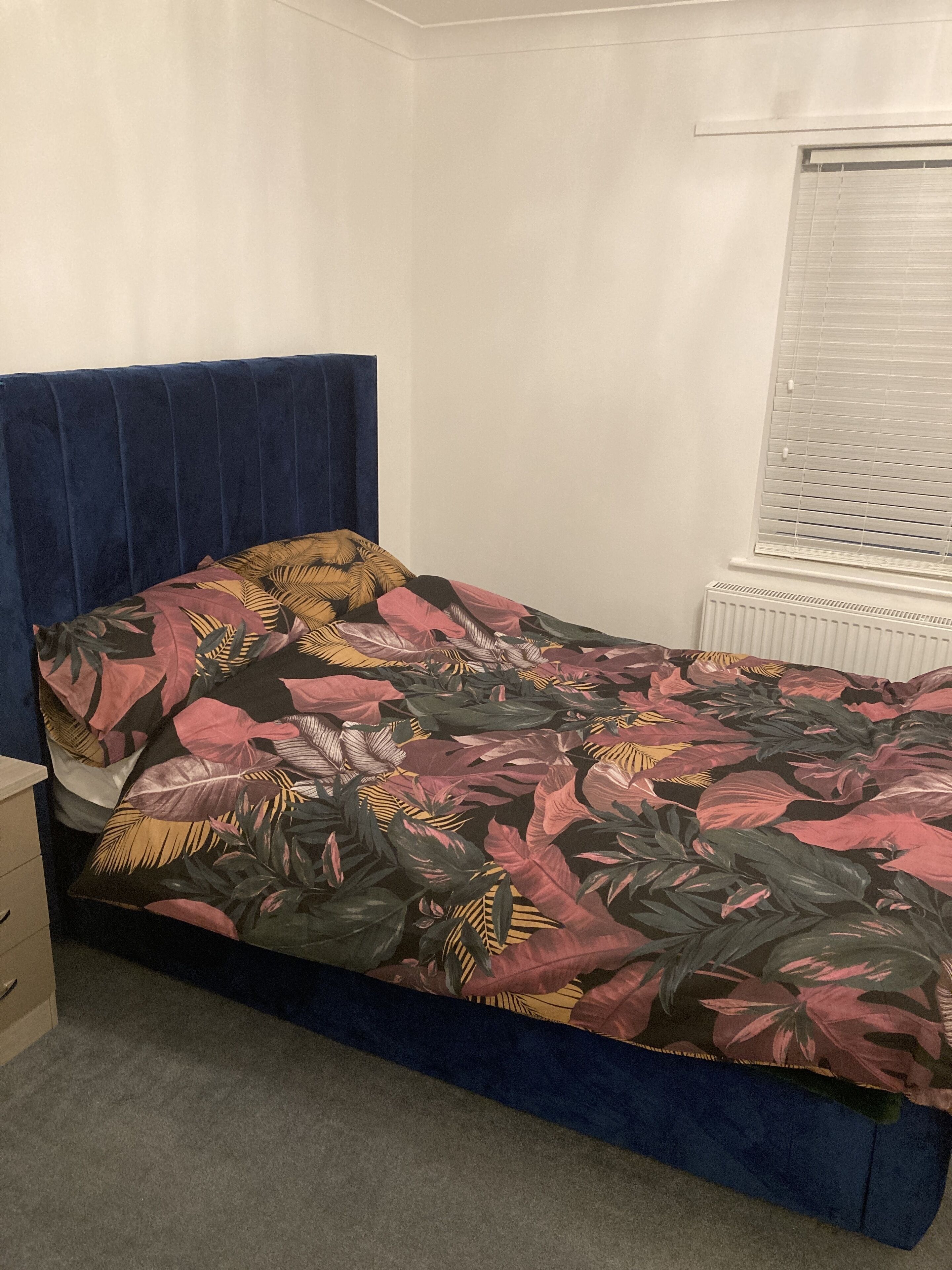 A bedroom with a bed, a blue headboard, a window with blinds, and a radiator.