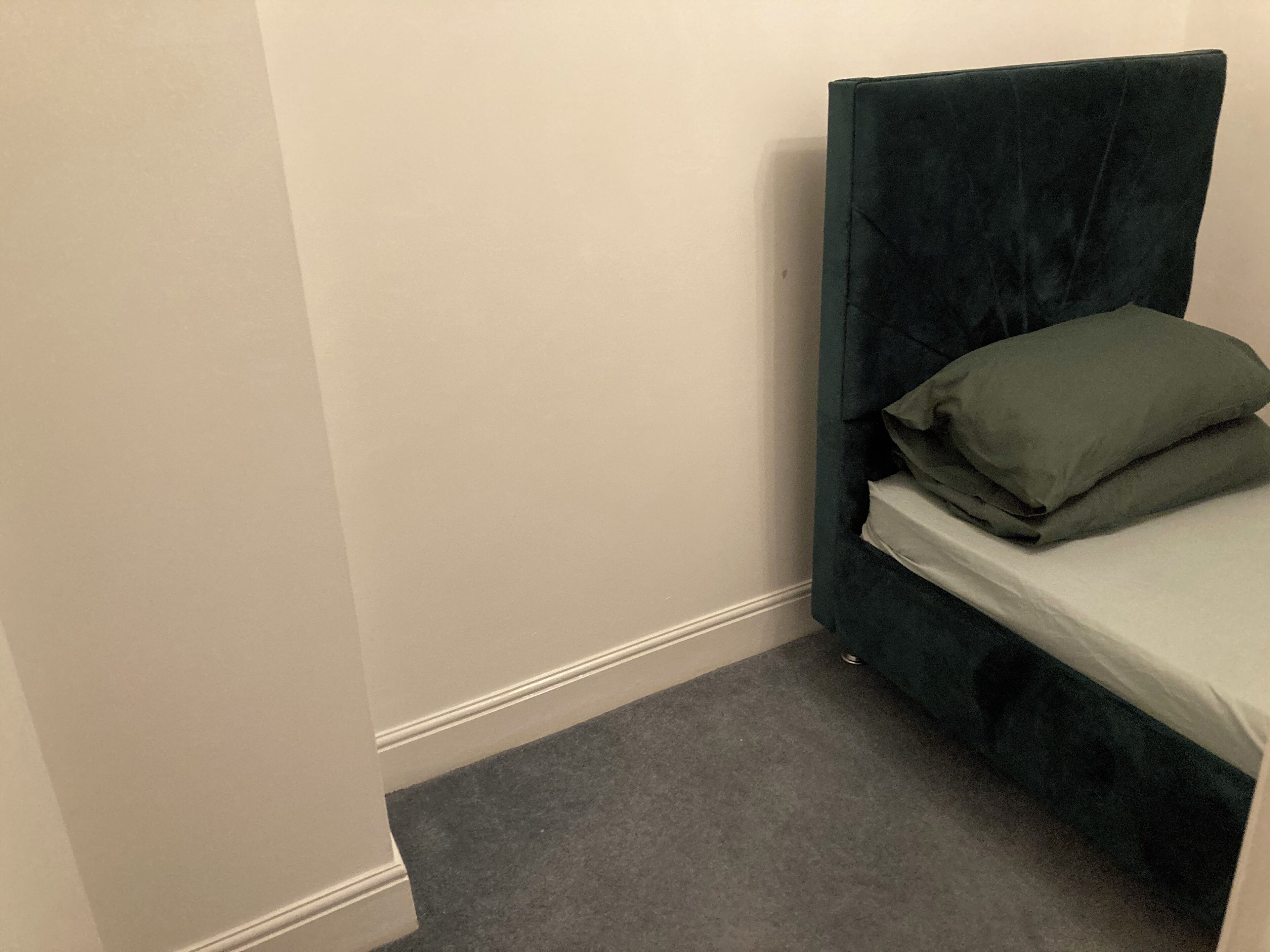 A small, dark green, single bed with a white mattress and a green pillow in a corner room with a white wall and a grey carpet.