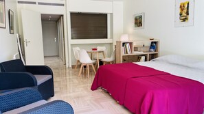 A room with a bed, a desk, two chairs, a bookshelf, and a door leading to another room.