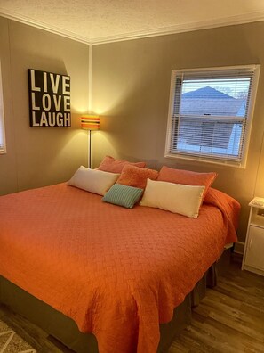 A bedroom with a large bed, a wall-mounted sign saying "LIVE LOVE LAUGH", a floor lamp, and a window with blinds.
