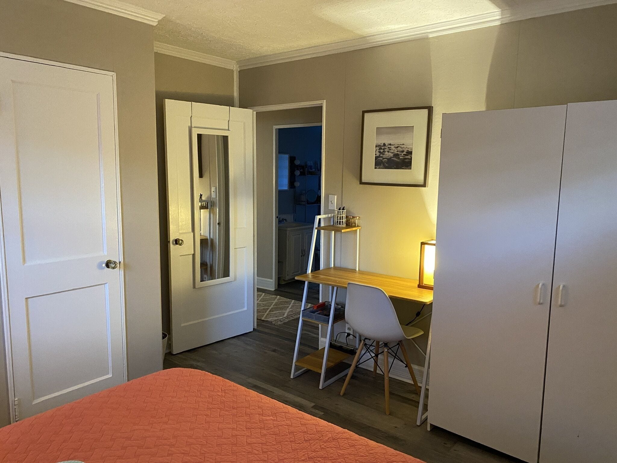A bedroom with a bed, a desk with a chair, a wardrobe, and a door leading to another room.
