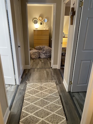 A hallway with a patterned rug, wooden flooring, and a bed with patterned bedding in the background.