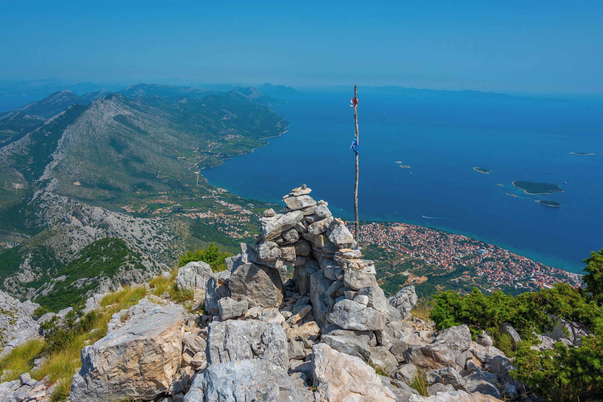 A rocky mountain peak with a flagpole, overlooking a coastal town and the sea.