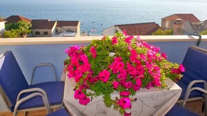 A balcony with a table covered by a tablecloth, surrounded by blue chairs, overlooking a body of water and distant hills. - Holiday apartment With sea view and terrace (Komarna)