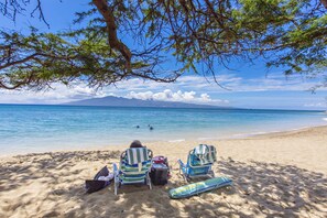 A beach with two striped beach chairs, a blue beach mat, and a bag on the sand.