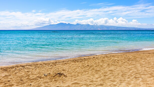Una playa con aguas turquesas cristalinas, una orilla arenosa y montañas lejanas bajo un cielo azul.