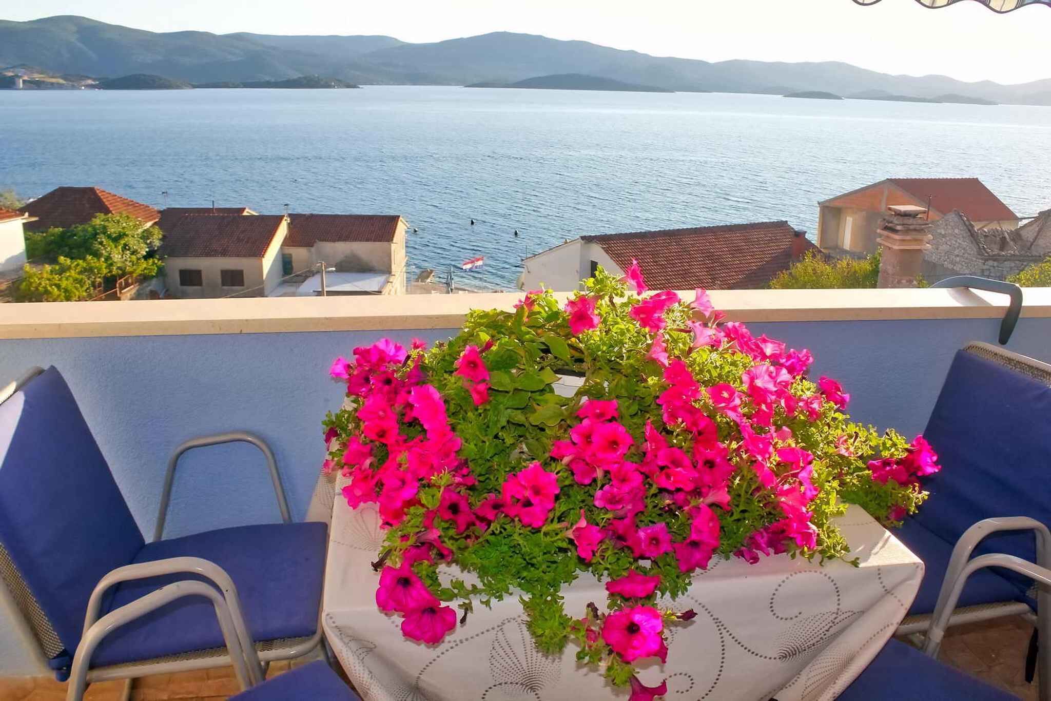 A balcony with a table covered by a tablecloth, surrounded by blue chairs, overlooking a body of water and distant hills.