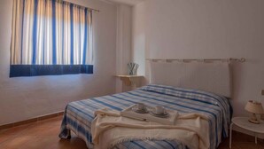 Ein Schlafzimmer mit Bett, Nachttisch, einem Fenster mit Vorhängen und einem kleinen Tisch mit Lampe.