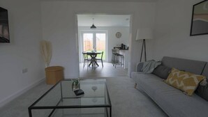 A living room with a glass coffee table, a grey sofa with a patterned throw pillow, a lamp, and a dining area with a table and chairs. - Beautiful 4-bed House 15 Mins to Cardiff & Newport (Cardiff)