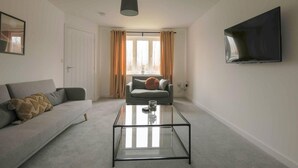A modern living room with a grey sofa, a glass coffee table, and a flat-screen TV mounted on the wall. - Beautiful 4-bed House 15 Mins to Cardiff & Newport (Cardiff)