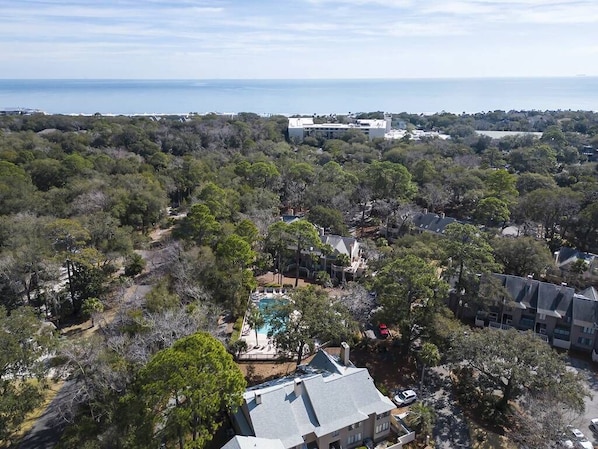 An aerial view of a residential area with a large house featuring a swimming pool, surrounded by dense trees and a view of the sea.