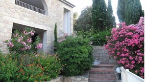 A stone building with a balcony, surrounded by various flowering plants and trees. - Maison Corse n°4 (Propriano)