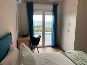 Una camera da letto con un letto, una scrivania e una sedia. Si apre una finestra con tende che dà su un panorama esterno.