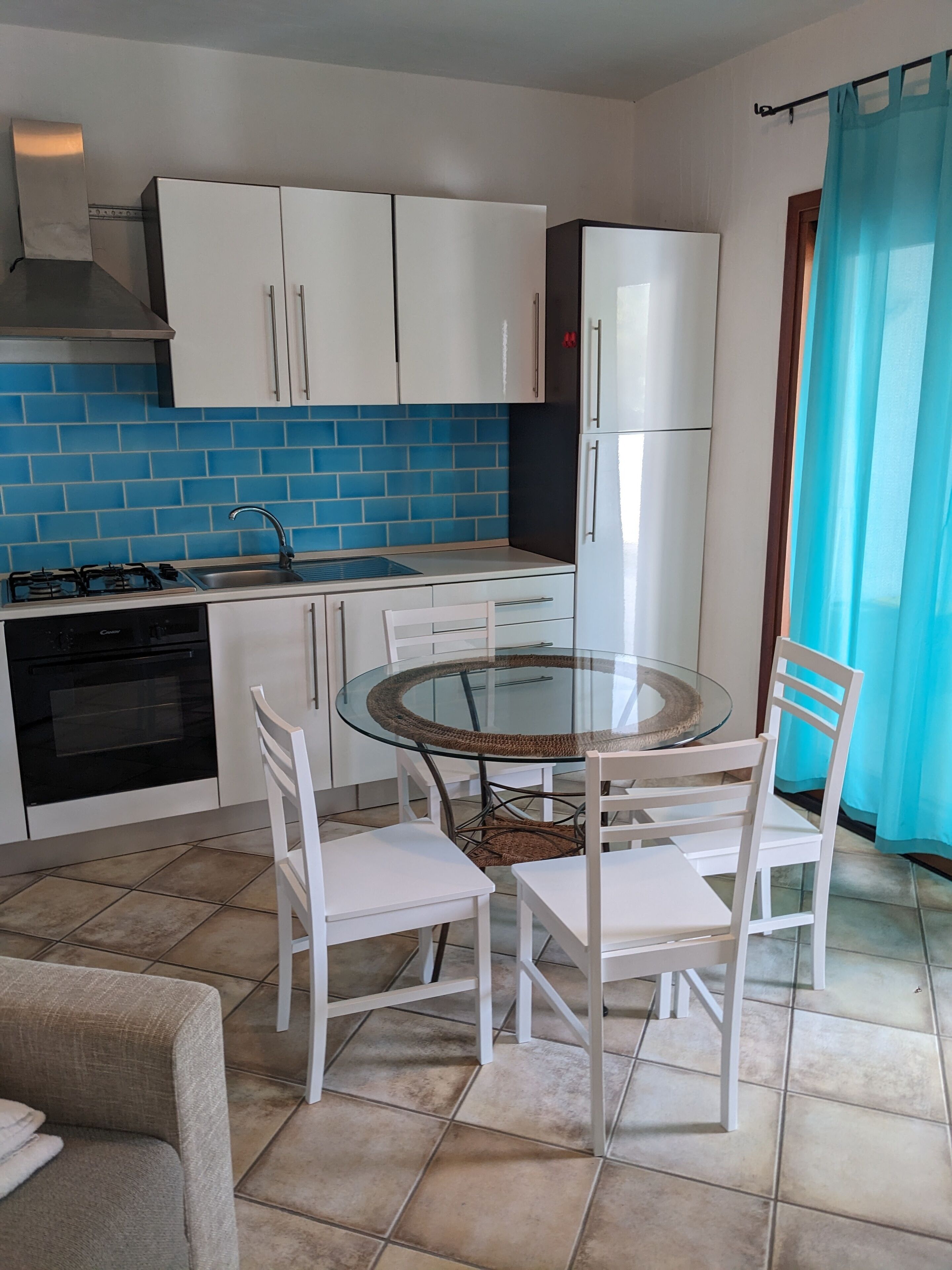 A compact kitchen with white cabinets, a blue-tiled backsplash, a gas stove, and a refrigerator. There is a round glass table with white chairs.