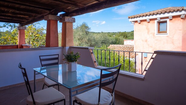 A balcony with a glass table and chairs, overlooking a residential area with trees and a building. - il Girasole (Golfo Aranci)