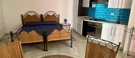 Un dormitorio compacto con una cama, una mesita de noche pequeña y una silla. Junto a él, una kitchenette con estufa, fregadero y armarios.