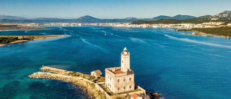 An aerial view of a coastal town with a prominent lighthouse, clear blue waters, and distant mountains.