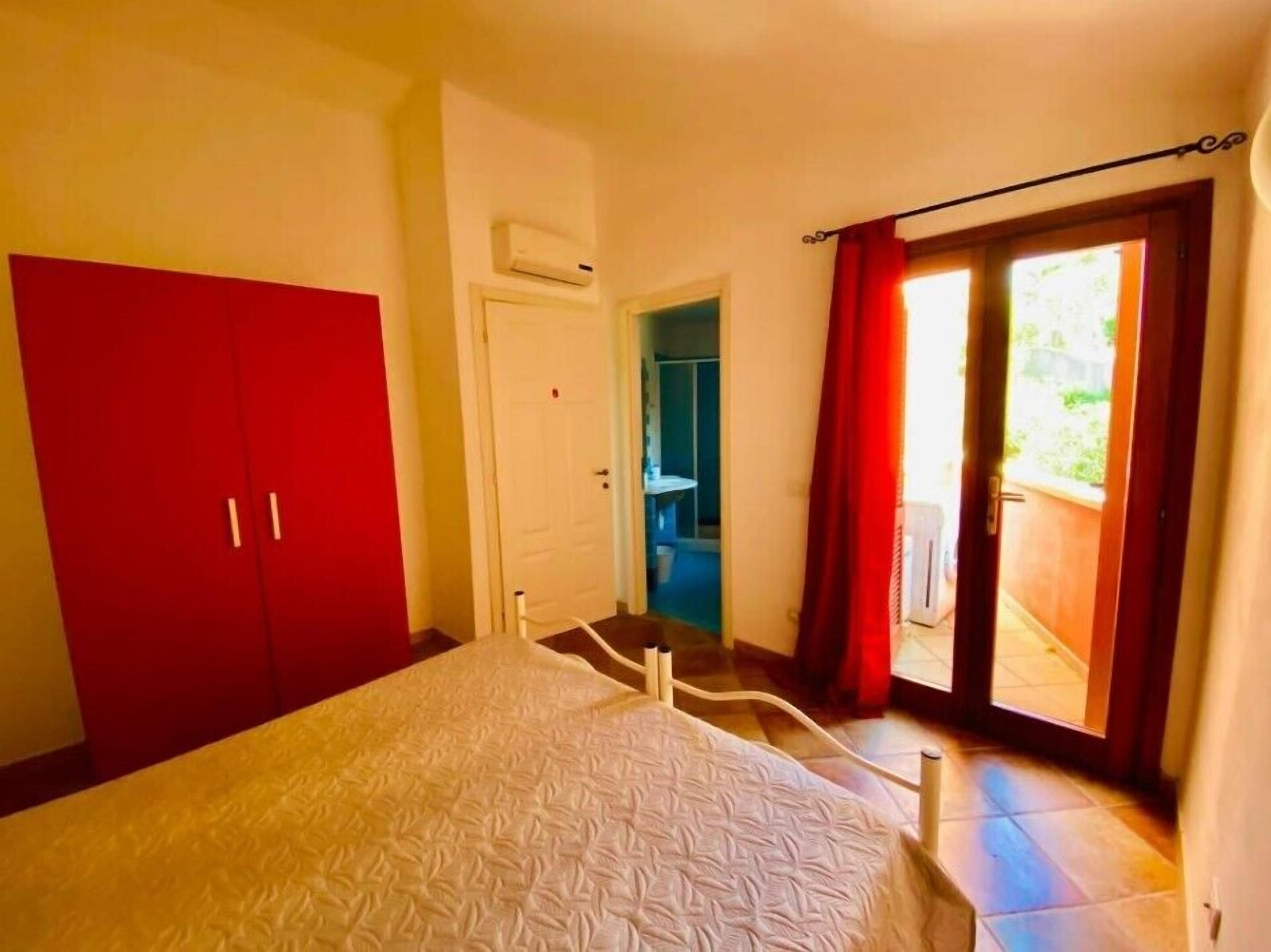 Un dormitorio con cama, un armario rojo, una puerta blanca y una puerta de cristal corredera que da a un balcón.