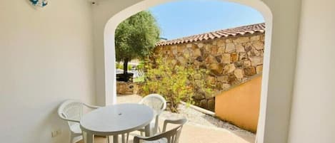 Un patio con una mesa redonda y dos sillas, un arco que da a un muro de piedra, y un árbol visible a través del arco.