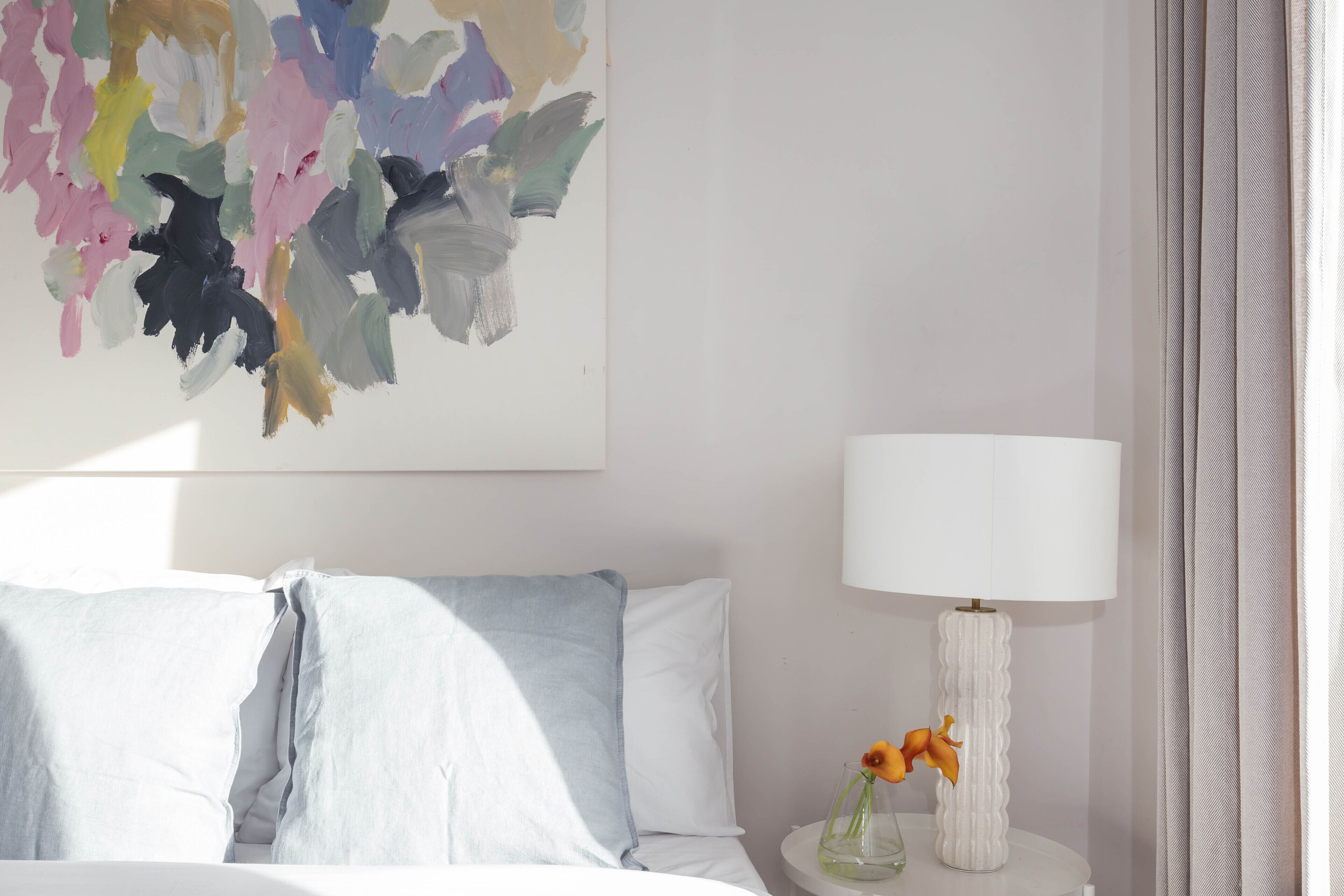 A bedroom with a bed, pillows, a bedside table with a lamp, and a wall painting.