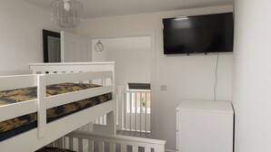 Una cama litera con ropa de cama de motivos florales, un armario blanco y un televisor montado en una habitación con paredes blancas y una puerta que da al exterior.
