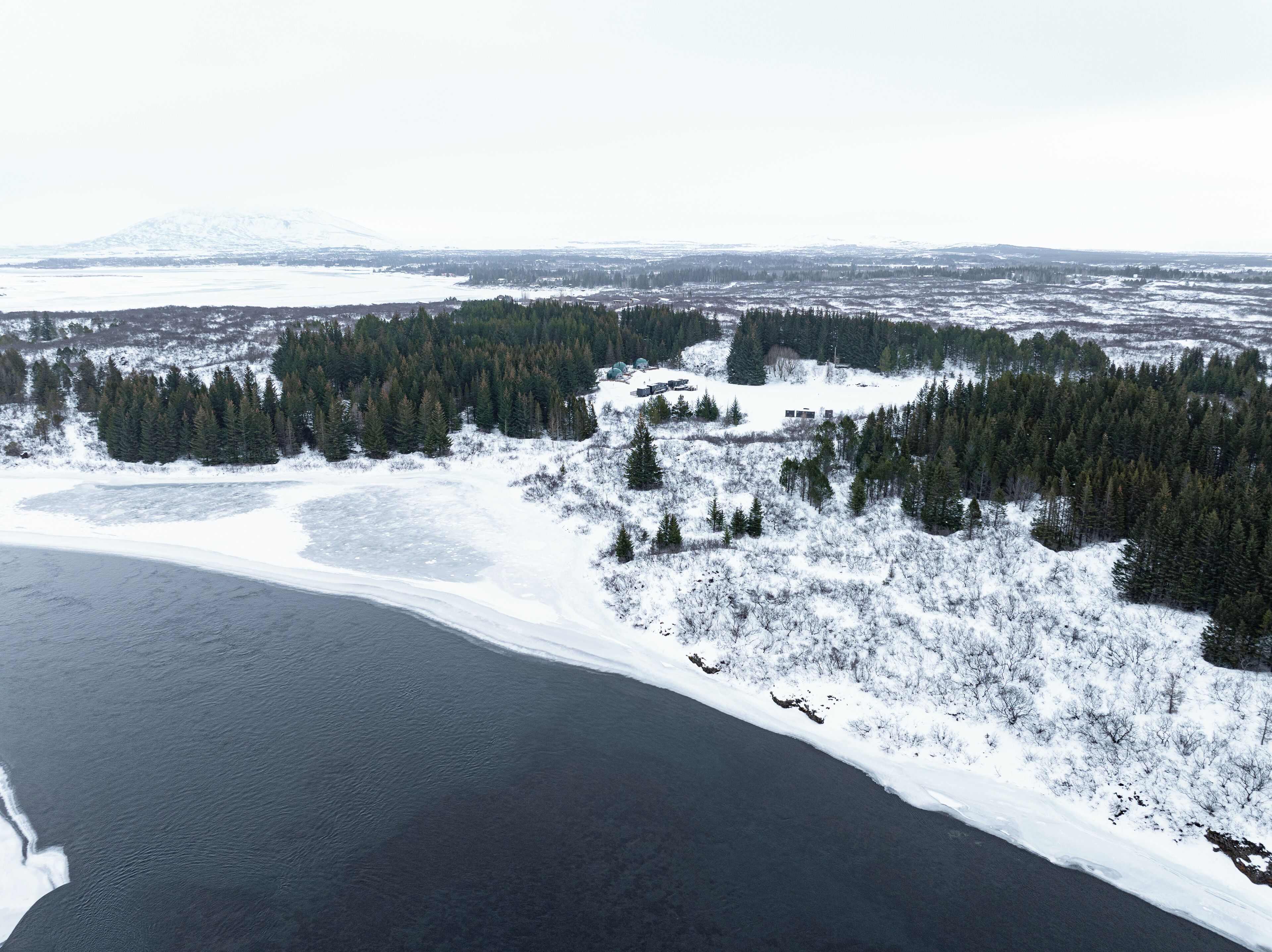 Une vue aérienne d’un paysage enneigé, avec un lac, une forêt dense et des montagnes lointaines.