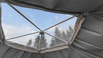 Une tente dotée d’une fenêtre transparente, offrant une vue sur les arbres et un ciel nuageux.