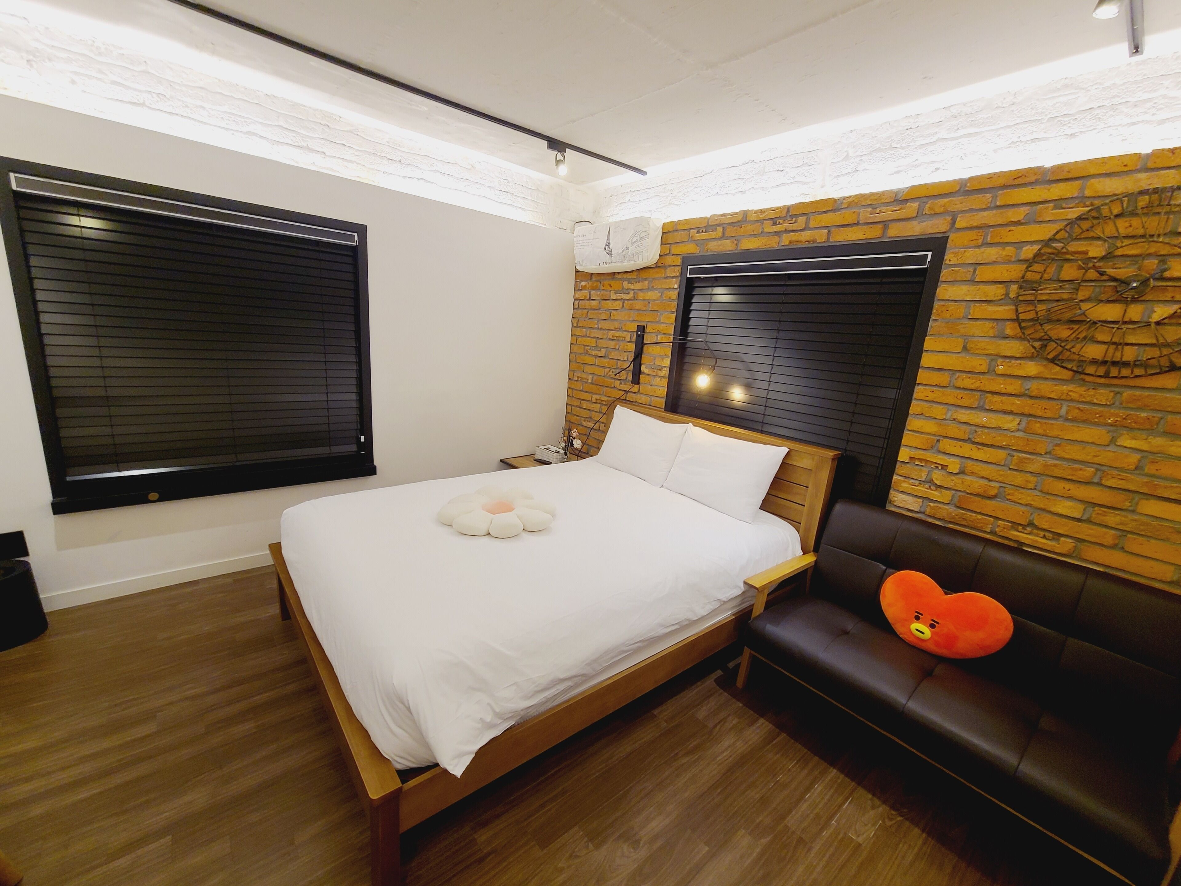 A bedroom with a wooden bed, two windows with blinds, a brick wall, a black leather couch, and a small table with a lamp.