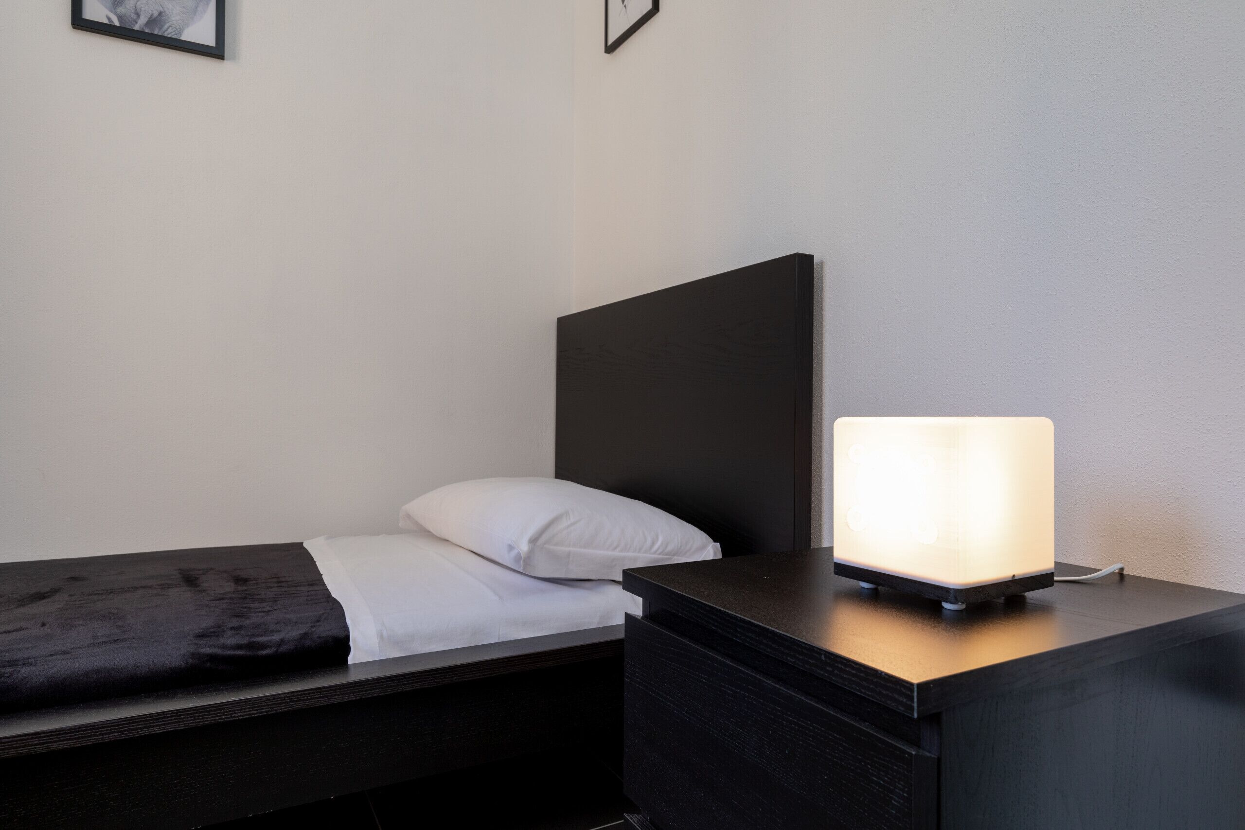Een minimalistisch ingerichte slaapkamer met een bed, een nachtkastje met een lamp en een aan de muur hangend schilderij.