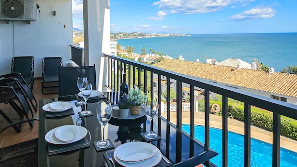 A balcony with a dining table set for two, overlooking a swimming pool and a coastal view.