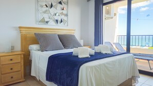 A hotel room with a bed, a wooden nightstand, a blue blanket, and a view of the sea through a balcony.