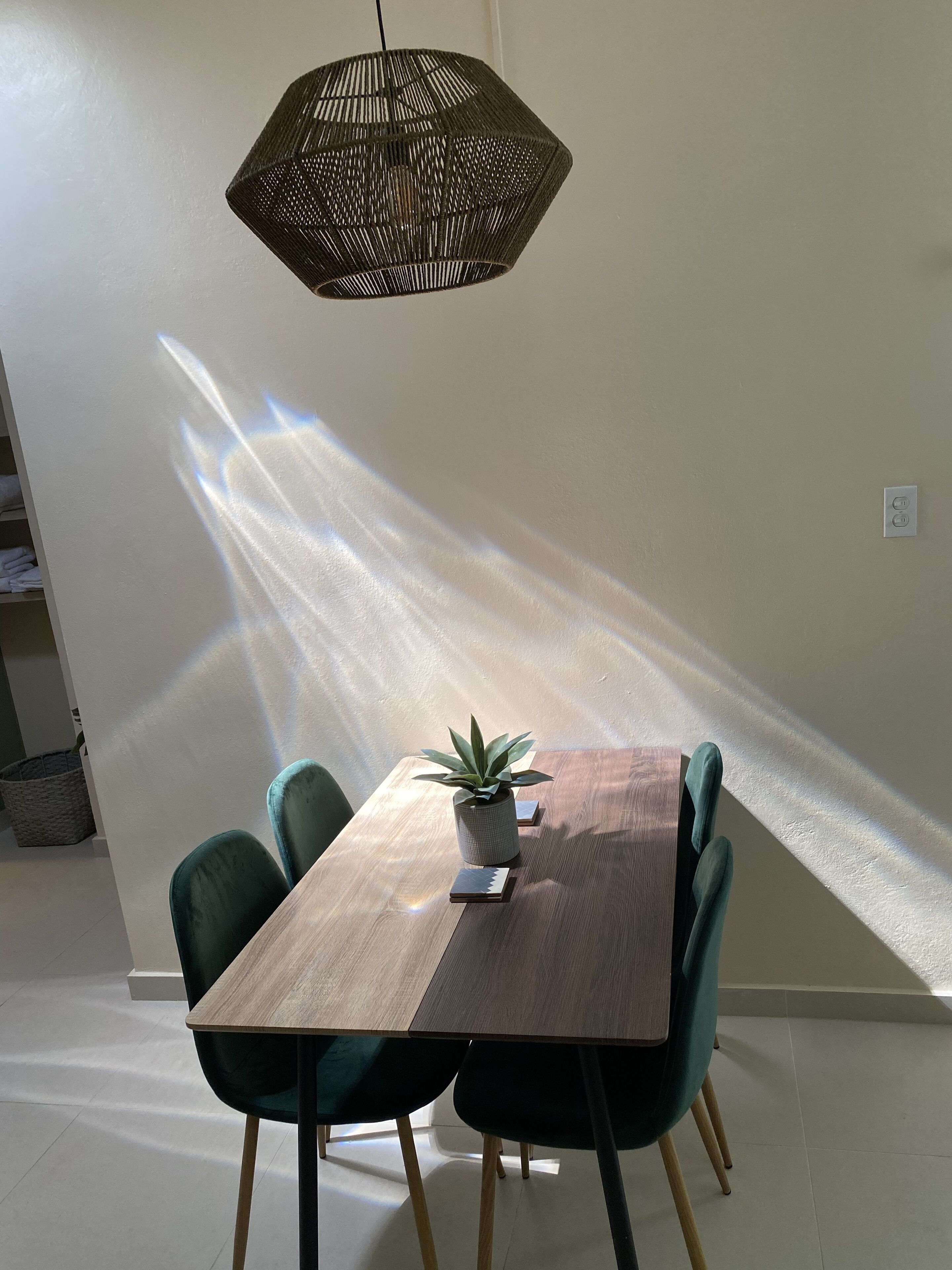 Un coin repas avec une table en bois, deux chaises vertes, une plante en pot et une suspension au design géométrique.