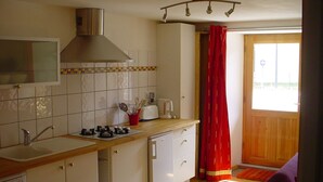 Pieni, käytännöllinen keittiö, jossa on pesuallas, liesi ja kaapit. Ikkunassa on punainen verho, ja puiset ovet johtavat ulos.