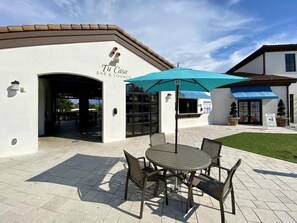 Eine Terrasse im Freien mit Tisch und Stühlen, einem blauen Sonnenschirm und einem Gebäude mit der Aufschrift 'Tu Casa Bar & Lounge'.