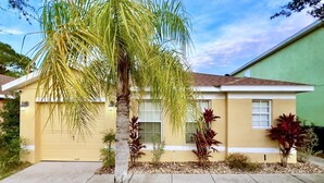 A single-story house with a yellow exterior, a garage door, and a small front yard with a palm tree and red-leafed plants. - Terrific Value 4bd Pool Gm Spa Hm Sandy Ridge-640b 4 Bedroom Home by RedAwning (Davenport)