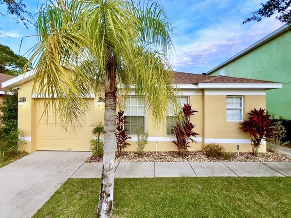 A single-story house with a yellow exterior, a garage door, and a small front yard with a palm tree and red-leafed plants.