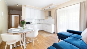 Cocina moderna con gabinetes blancos, microondas integrado y campana extractora de acero inoxidable. Hay un comedor con mesa de vidrio y sillas blancas, y un sofá azul en la sala de estar.